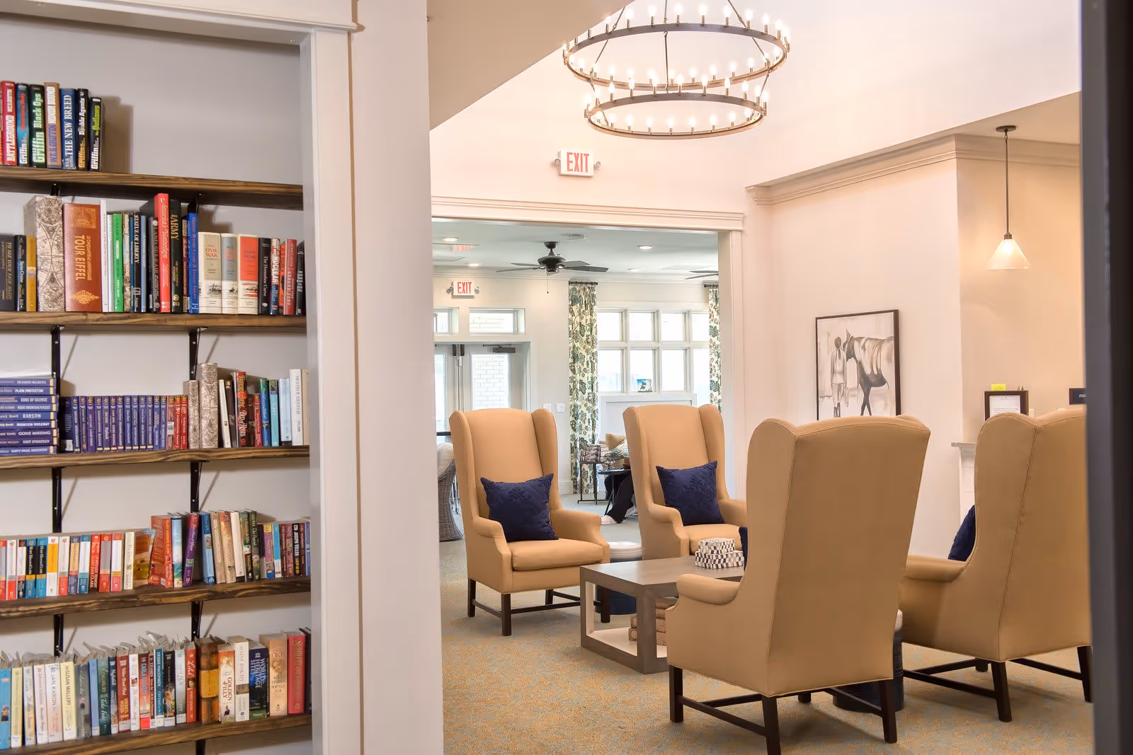 A cozy senior living common area with four beige armchairs arranged around a wooden coffee table. The chairs have dark blue pillows. To the left, there is a bookshelf filled with various books. The room is well-lit with natural light coming through large windows in the background and a modern chandelier hanging from the ceiling.