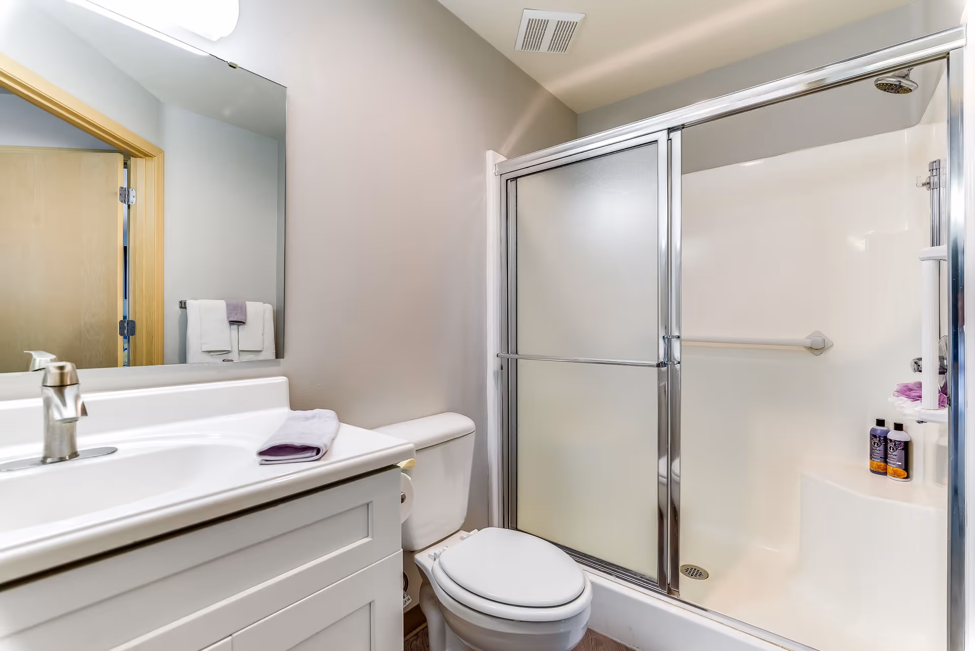 A clean and modern bathroom featuring a white vanity with a sink and faucet, a toilet, and a shower with frosted sliding glass doors. There are two bottles of shower products and a purple bath sponge inside the shower. A towel is folded on the vanity, and two towels hang on a rack reflected in the mirror above the sink.