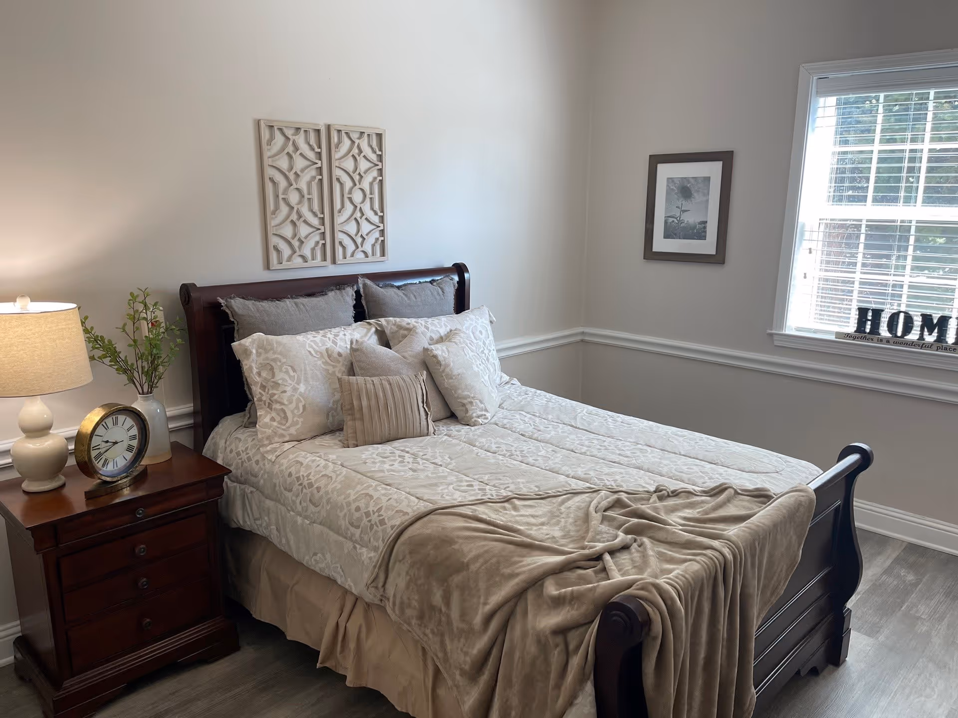 Bright bedroom featuring a wooden sleigh bed dressed in neutral bedding, a nightstand with a lamp and clock, wall art and a window.