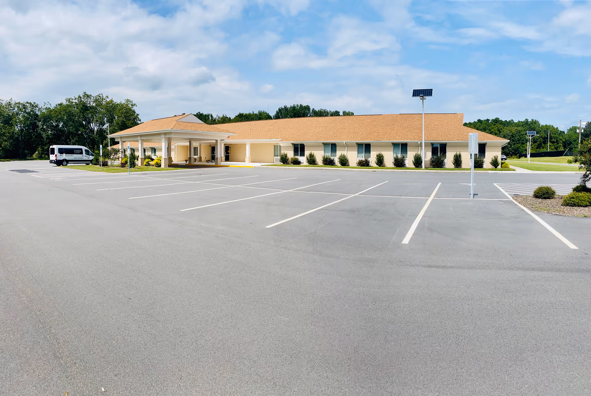 Wide-angle view of a single-story beige senior living building with a covered entrance, adjacent van, and a large empty parking lot under a partly cloudy sky.