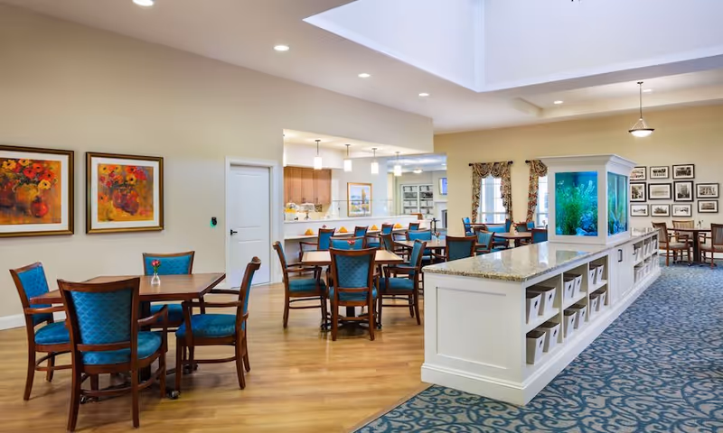 Bright senior living dining area with wooden tables and teal upholstered chairs, a long white buffet with a built-in aquarium, and pendant lighting.