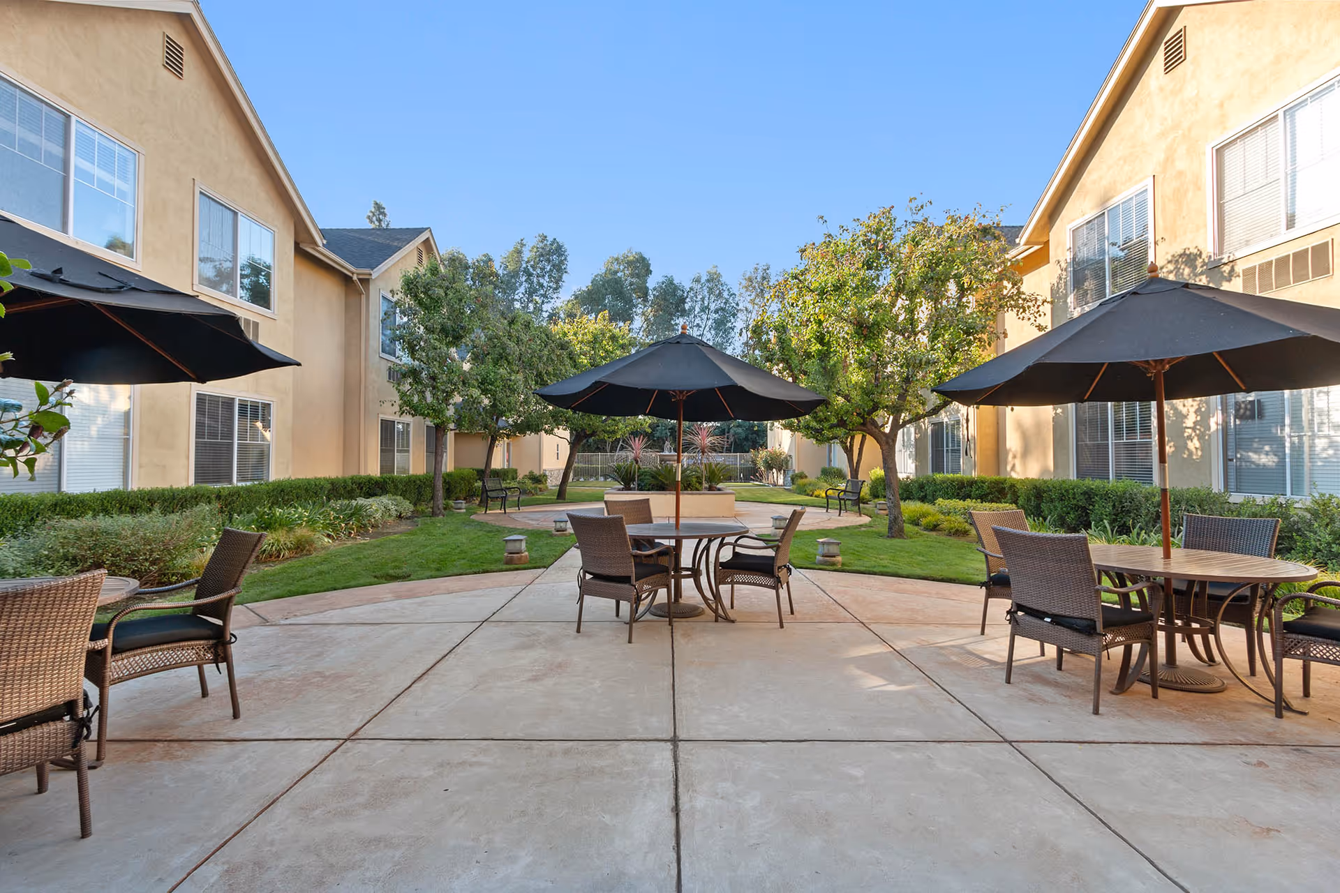 Courtyard patio between two residential buildings with tables, chairs, and black umbrellas on a paved area.