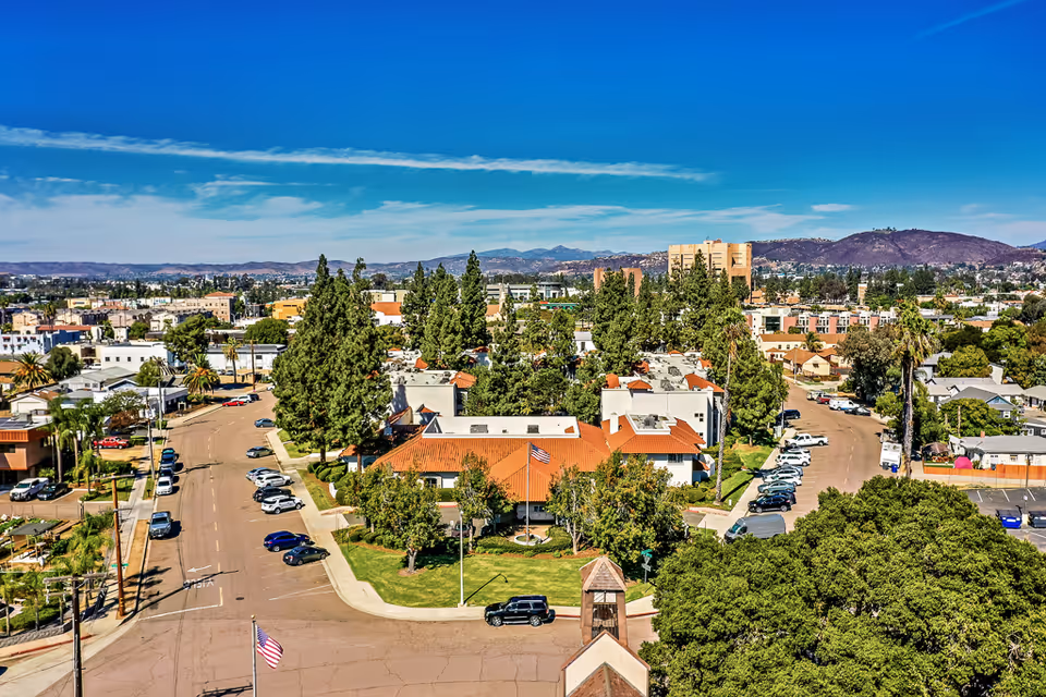 Aerial view of a residential campus with red-tile-roof buildings, trees, parking areas and surrounding neighborhood under a clear blue sky.