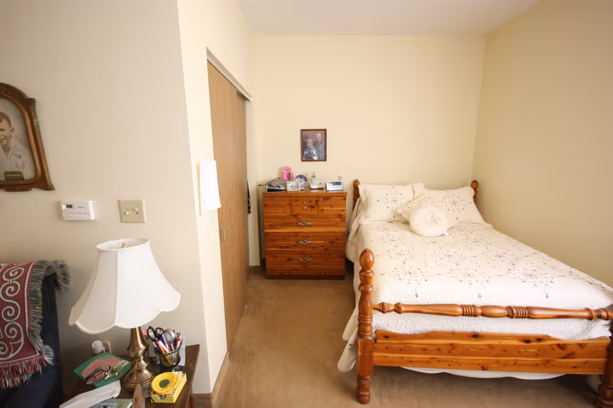 A small bedroom with a wooden bed covered in a white quilt, a dresser against the back wall, and a nightstand with a lamp and personal items.