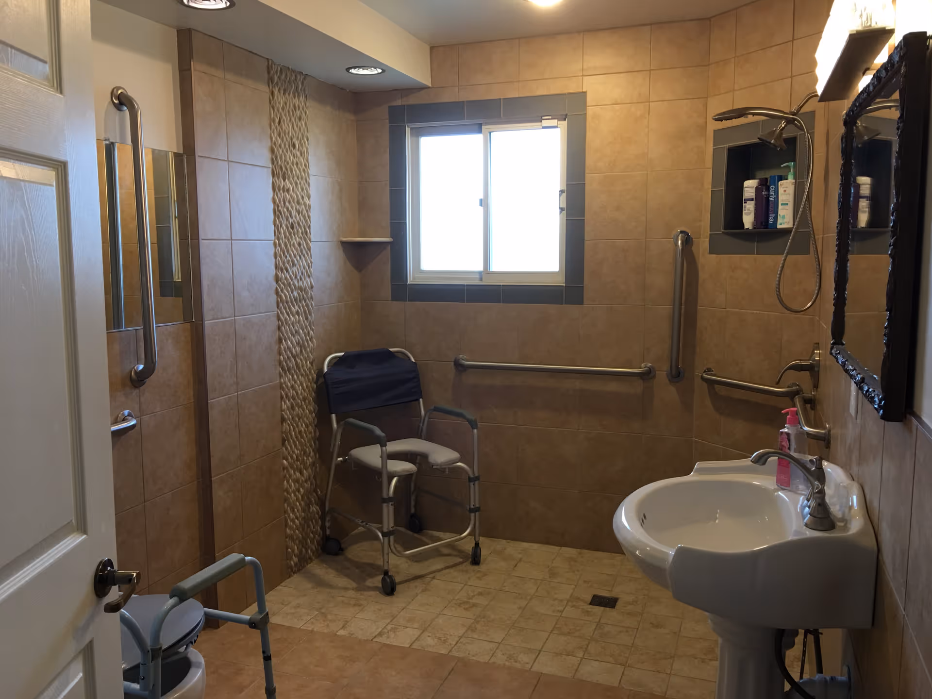A bathroom with beige tiled walls and floor, featuring a shower area with a shower chair, grab bars on the walls, a pedestal sink with a mirror above it, and a window letting in natural light.