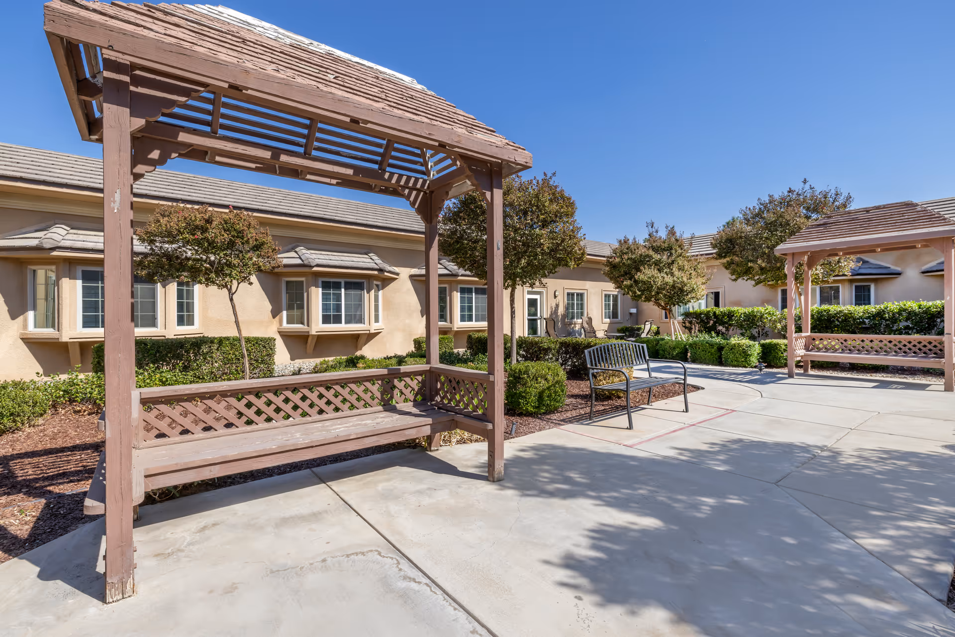Outdoor courtyard area at Brookdale Riverwalk featuring two wooden pergola benches, a metal bench, small trees, shrubs, and a beige building with multiple windows in the background under a clear blue sky.