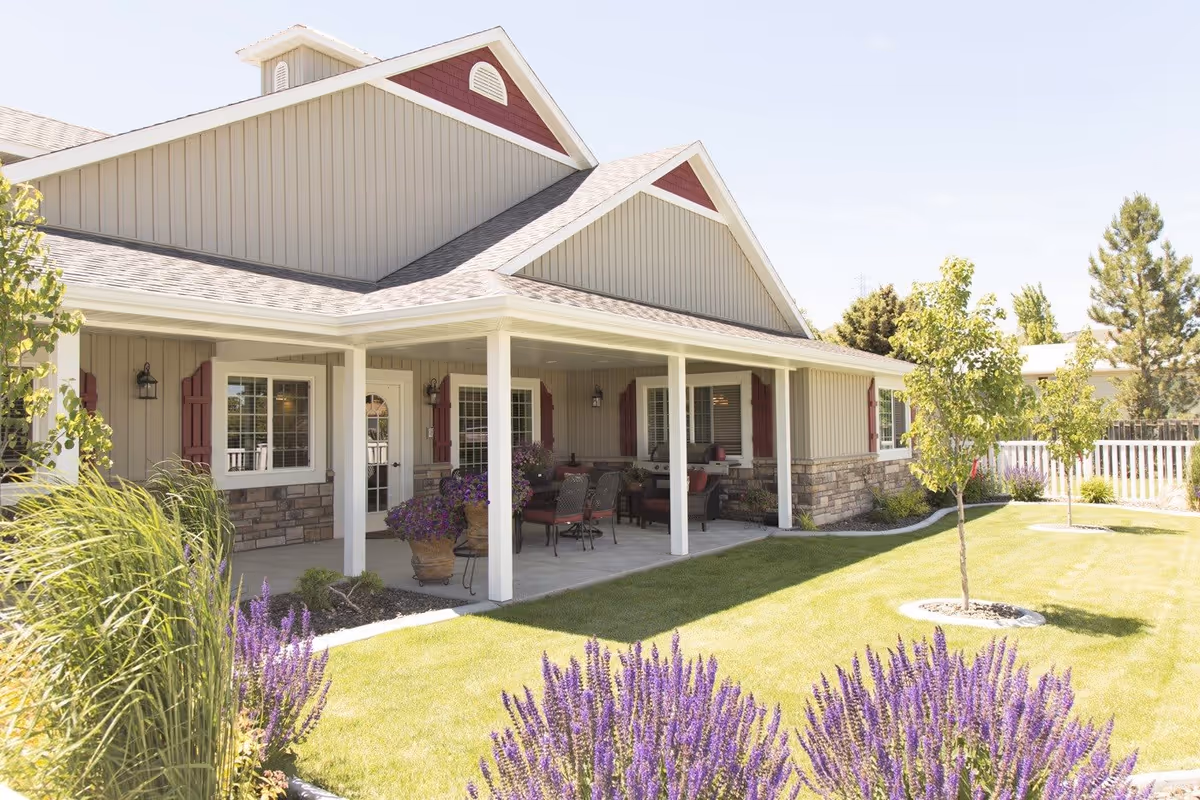 Exterior view of a single-story building with beige siding and red shutters, featuring a covered porch with outdoor seating. The foreground shows a well-maintained lawn with small trees and purple flowering plants under a clear sky.