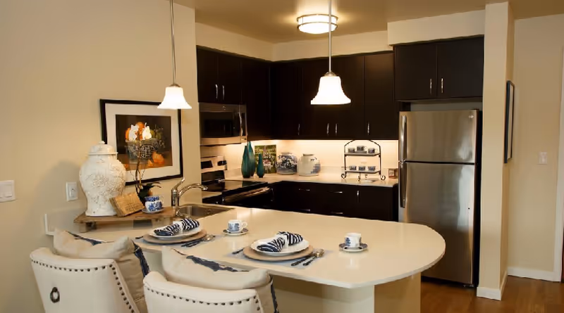 Modern kitchen with dark wood cabinets, stainless steel refrigerator and microwave, and a white countertop island set with plates, cups, and napkins. Two cushioned chairs are placed at the island, and decorative items including a large white vase and framed artwork are visible.