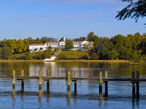 White multi-story building on a tree-lined hill across a calm body of water with wooden docks in the foreground.