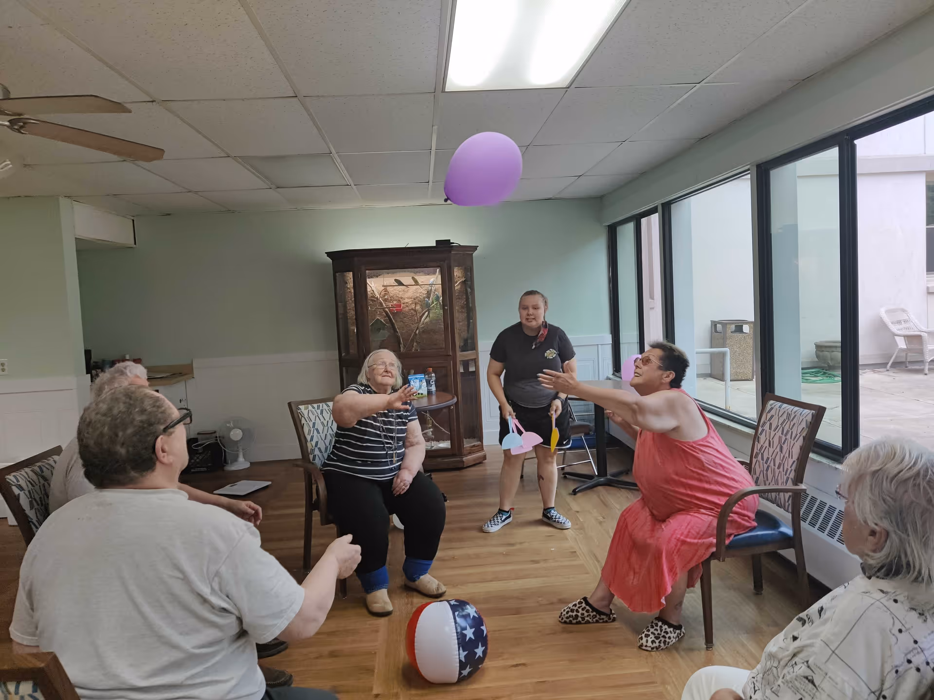A group of elderly people sitting in chairs arranged in a circle in a room with large windows. They are playing with a purple balloon and a beach ball on the floor. A younger woman stands in the background holding colorful paddles, facilitating the activity. The room has light green walls, wooden flooring, and a ceiling fan.