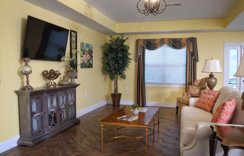 A bright living room with yellow walls, a sofa and armchair, wall-mounted TV, decorative cabinet, coffee table, and a potted plant.