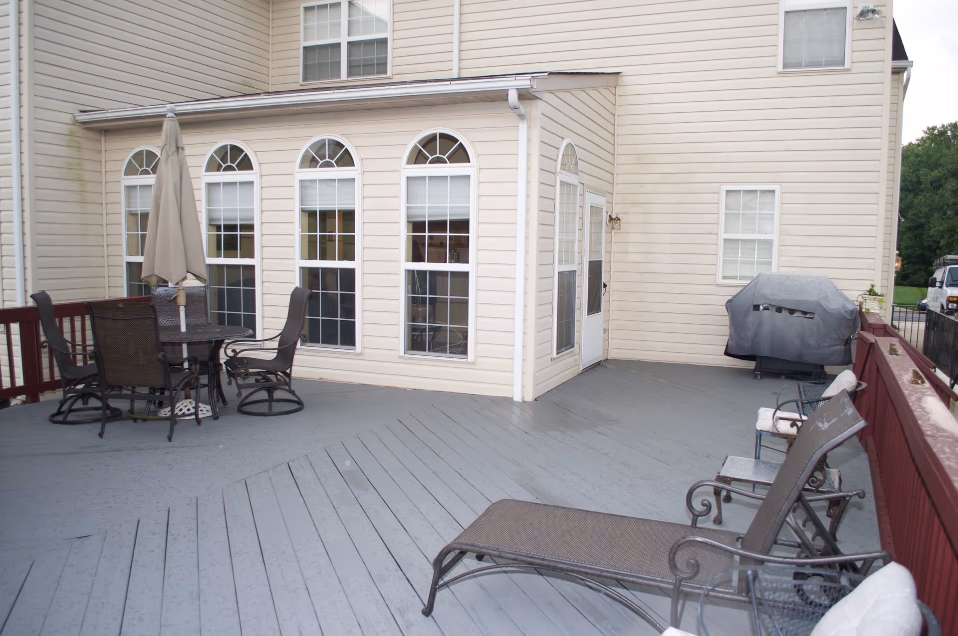 Outdoor patio area with gray wooden decking attached to a beige building. The patio features a round table with four chairs and a closed umbrella, a covered grill, two cushioned benches, and a lounge chair. The building has multiple windows with white frames and a white door leading to the patio.