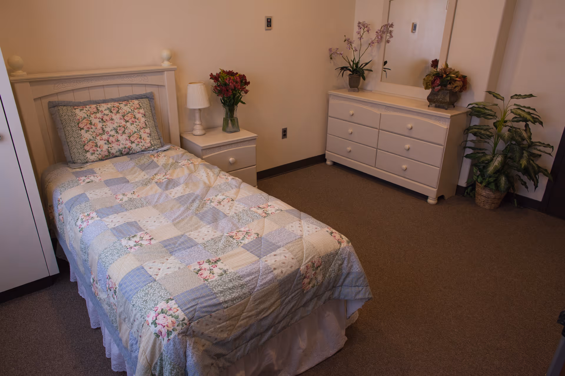 A small bedroom with a single bed covered in a patchwork quilt with floral patterns. Next to the bed is a white nightstand with a lamp and a vase of flowers. Across from the bed is a white dresser with six drawers, a large mirror, and two potted plants on top and beside it. The room has beige walls and carpeted floor.