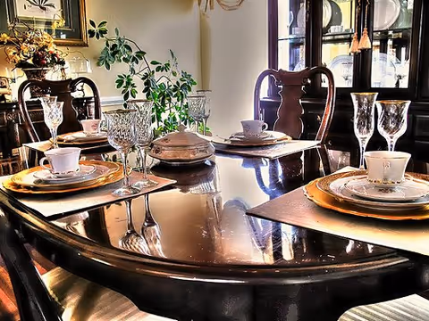 A polished wooden dining table set with china, crystal glassware, and place settings in a warmly lit dining room with chairs and a china cabinet.