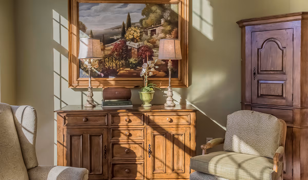 A cozy living room area with wooden furniture including a cabinet and a sideboard. Two table lamps with beige shades sit on the sideboard, which also holds a green potted plant and a book. A large framed painting depicting a scenic landscape hangs on the wall above the sideboard. Two upholstered armchairs with light patterned fabric are positioned near the furniture. Sunlight streams through a window, casting shadows on the walls and furniture.