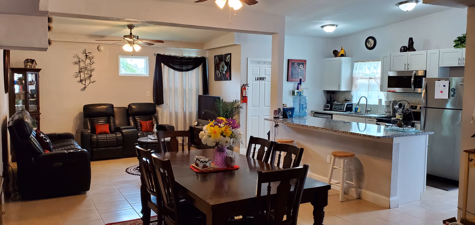 Interior view of a senior living facility showing a combined living and dining area with a kitchen. The living area has black leather recliners and a TV, the dining area has a wooden table with chairs and a flower centerpiece, and the kitchen features white cabinets, stainless steel appliances, and a granite countertop with bar stools.