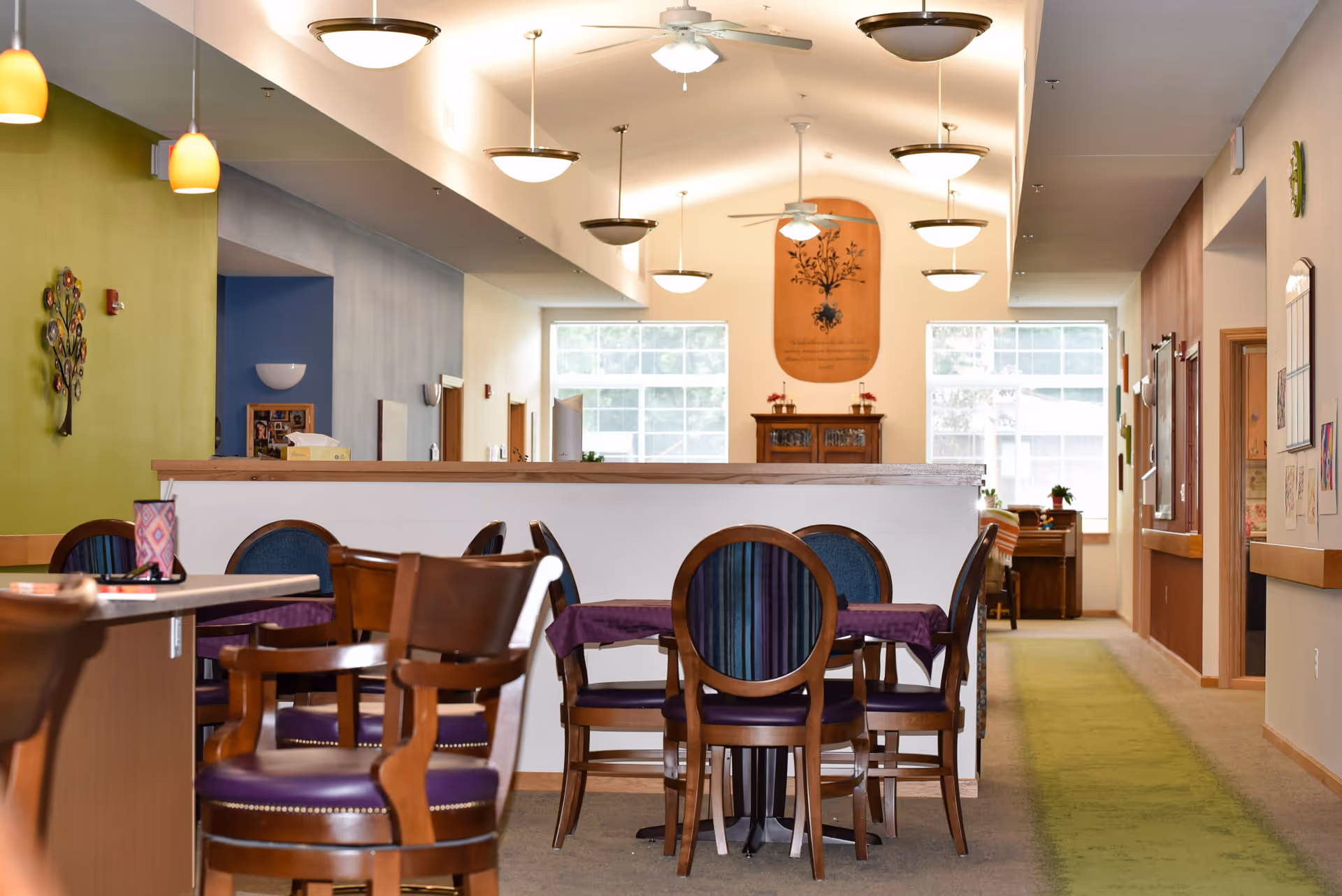 Interior view of a senior living facility dining area with wooden chairs and tables covered with purple tablecloths. The room features a green accent wall with decorative art, pendant and ceiling lights, and large windows letting in natural light. A hallway with carpet runs alongside the dining area.