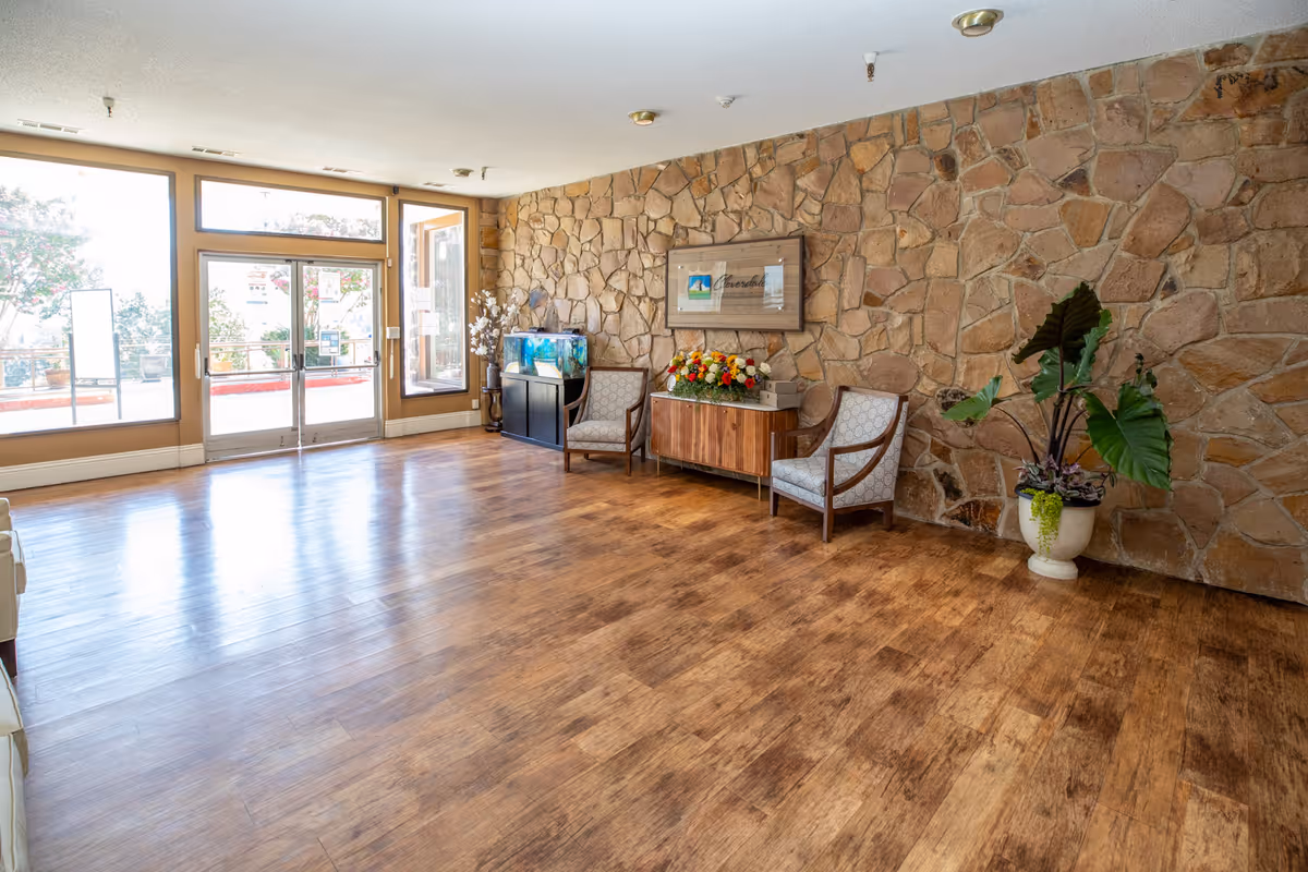 A spacious room with wooden flooring and a stone accent wall. The room features two upholstered chairs, a wooden cabinet with a flower arrangement on top, a fish tank, and a large potted plant. Large windows and glass doors allow natural light to fill the space.