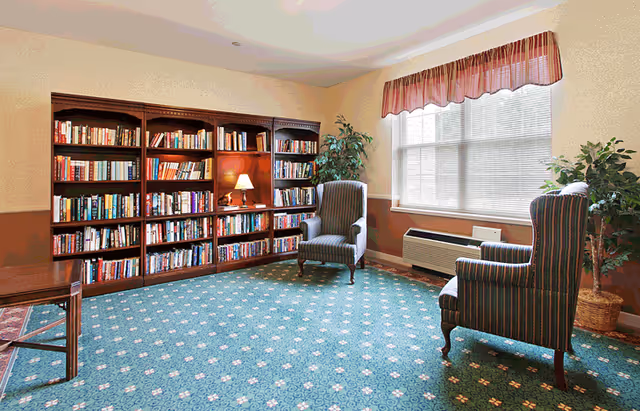 Sunlit common room with bookshelves, two upholstered armchairs, a table lamp, potted plants, and a window.