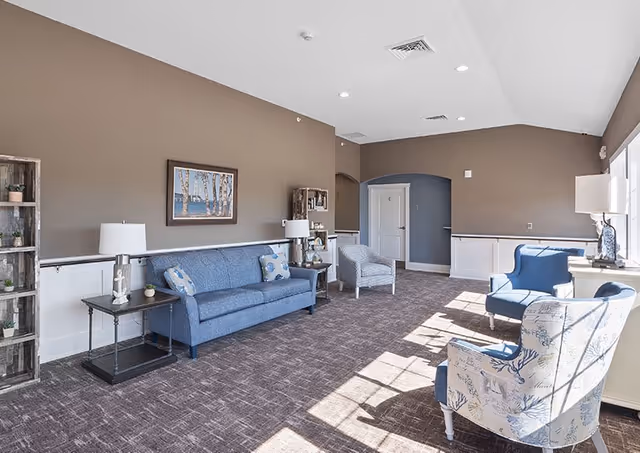 A bright and spacious living room with brown walls and carpeted floor. The room features a blue sofa with patterned pillows, two blue armchairs, and one white armchair with blue floral patterns. There are side tables with lamps and decorative items, a framed picture on the wall, and large windows letting in natural light.