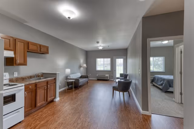 Interior view of a senior living apartment at Cedarhurst of Breese featuring a small kitchen area with wooden cabinets and a white stove on the left, a living room with a couch, two chairs, and a small table with a lamp, and a bedroom visible through an open doorway on the right. The room has wood flooring, neutral-colored walls, and windows letting in natural light.
