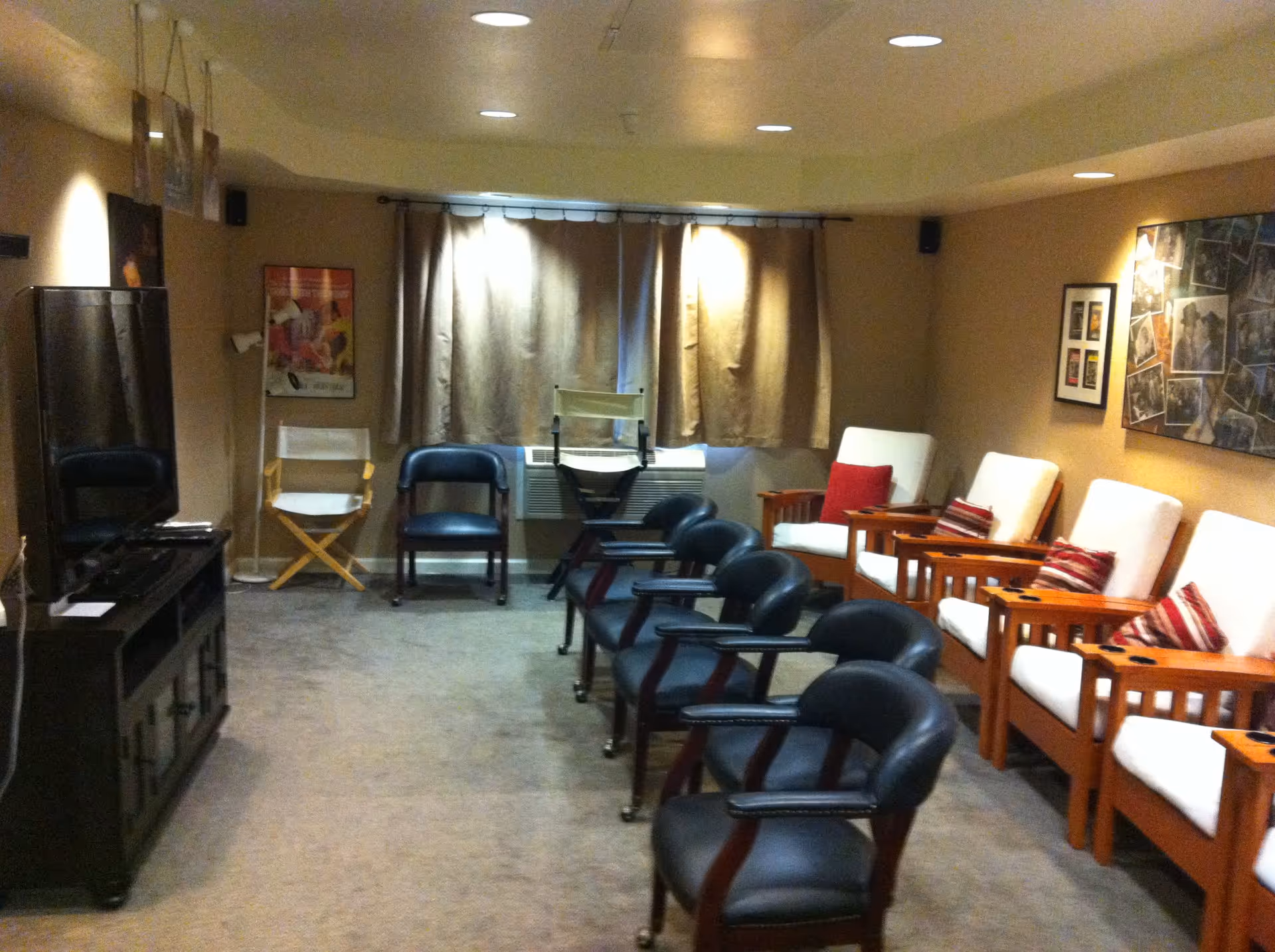 A cozy common room with two rows of chairs facing a large flat-screen TV on a wooden stand. The left row has black chairs with armrests, and the right row has wooden armchairs with white cushions and red striped pillows. The room has beige walls, carpeted floor, a window with closed curtains, and framed pictures on the walls.