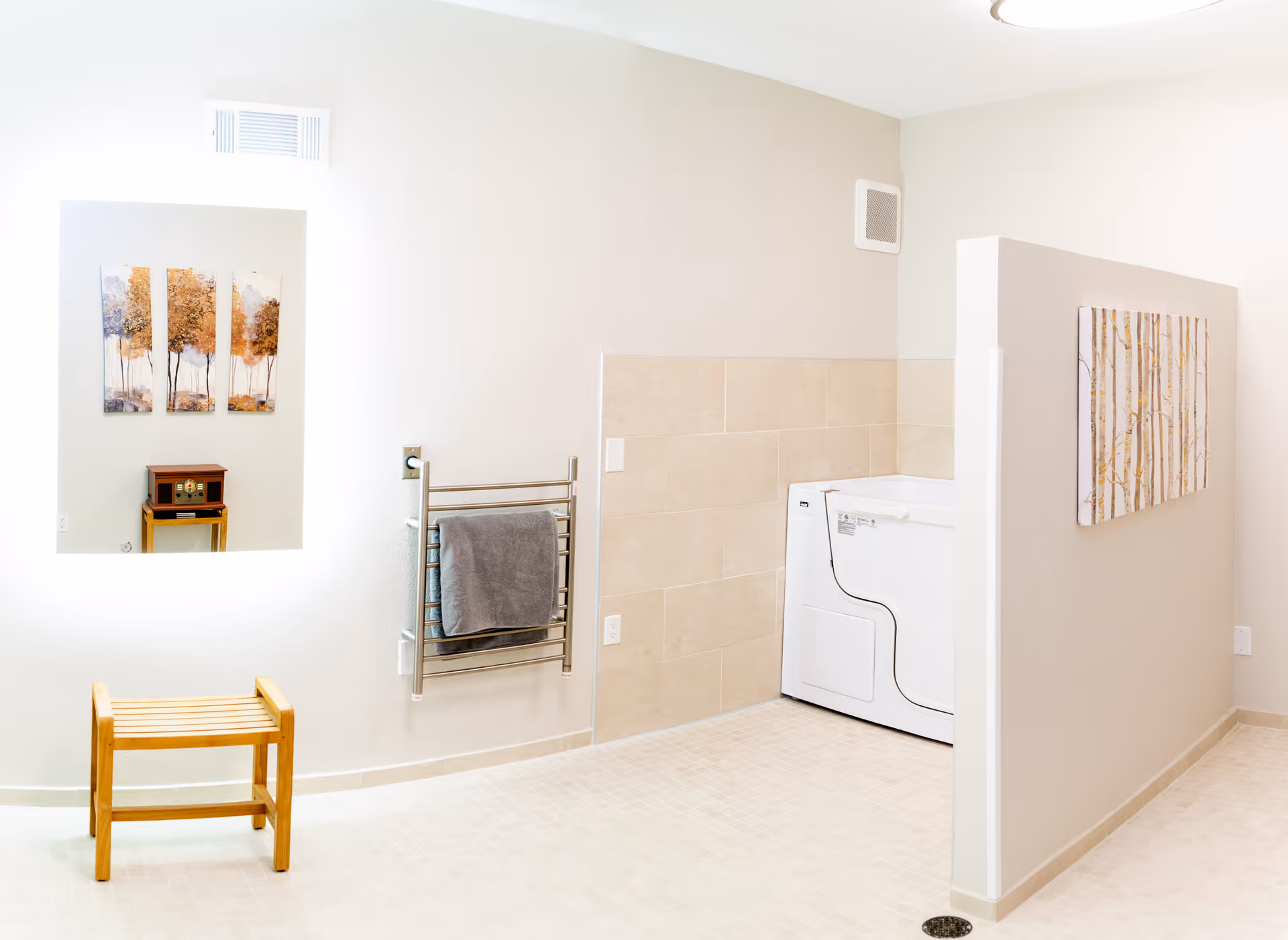 A bright, clean bathroom featuring a walk-in bathtub with safety rails, beige tiled walls, a towel rack with a gray towel, a wooden bench, and wall art depicting trees. A large mirror reflects a small table with a vintage radio and additional tree artwork.