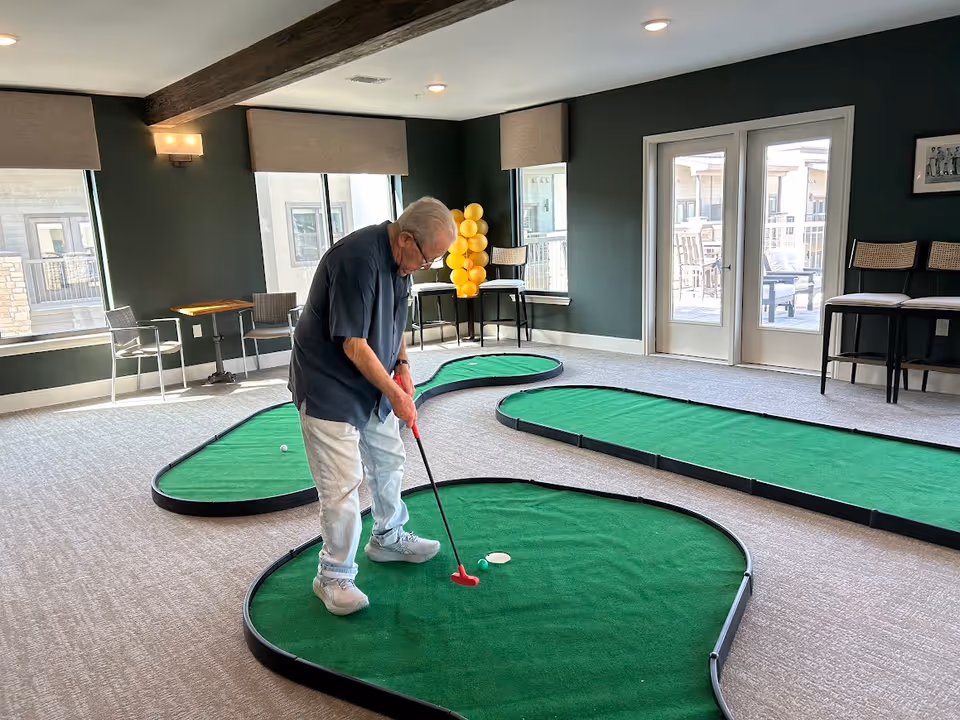 An elderly man playing indoor mini golf on a green putting surface inside a room with large windows and glass doors. The room has dark green walls, beige carpet, and several chairs along the walls. There is a cluster of yellow balloons near the windows.