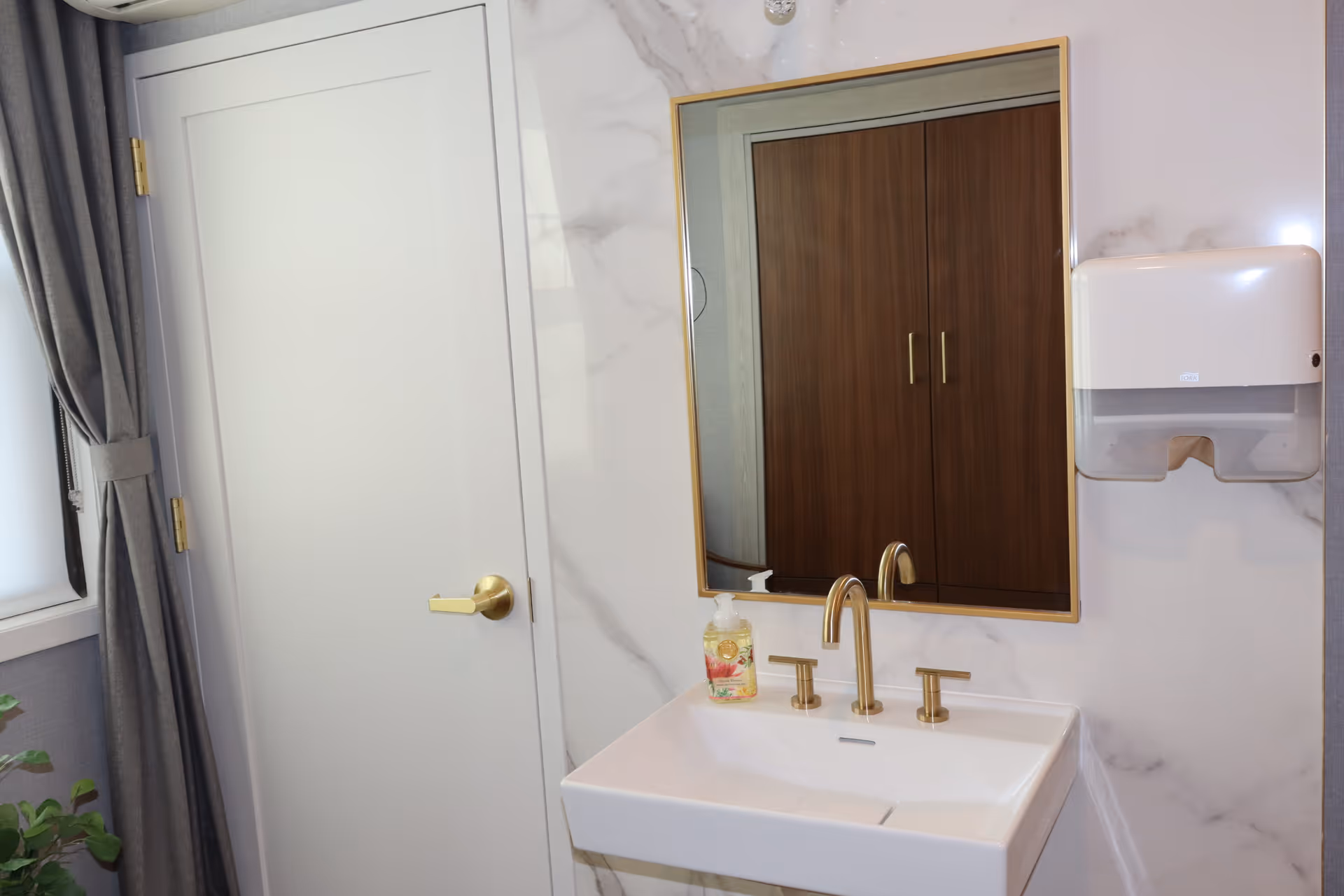 A clean bathroom sink area with a white rectangular sink, gold faucet and handles, a bottle of liquid soap, a wall-mounted paper towel dispenser, a large mirror with a gold frame reflecting a wooden cabinet, a white door with a gold handle, and a window with gray curtains.