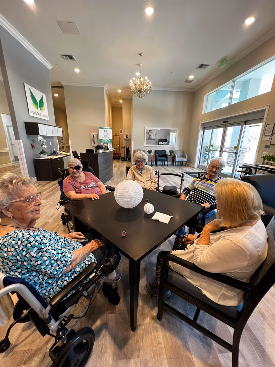 Five elderly women sitting around a square black table in a bright, spacious common area of a senior living facility. The room has light-colored walls, wood flooring, a chandelier, and large windows near the entrance door. Some women are in wheelchairs and others in chairs. The facility's reception desk and signage for Angels Senior Living are visible in the background.