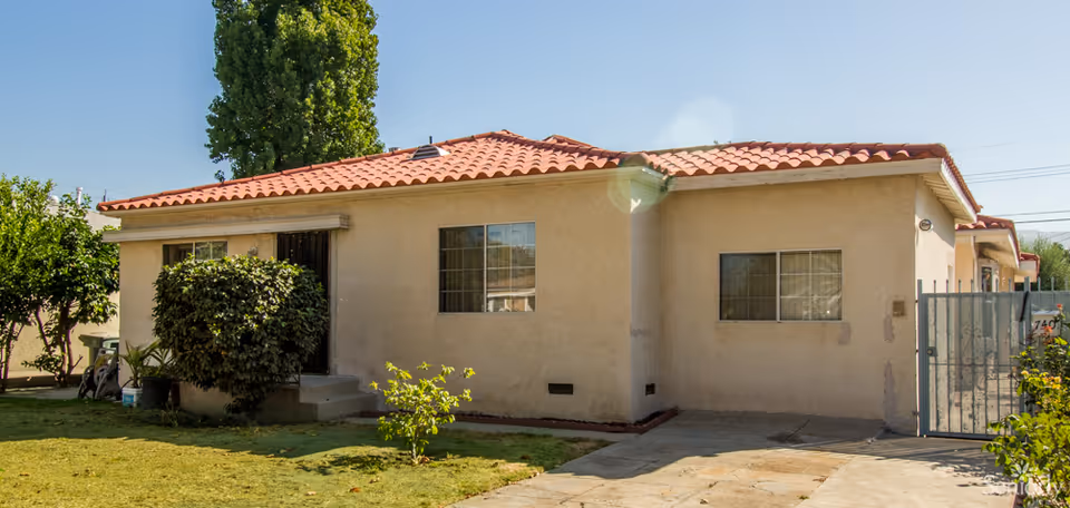 Single-story building with beige stucco walls and a red tile roof, surrounded by a small lawn with bushes and trees under a clear blue sky.