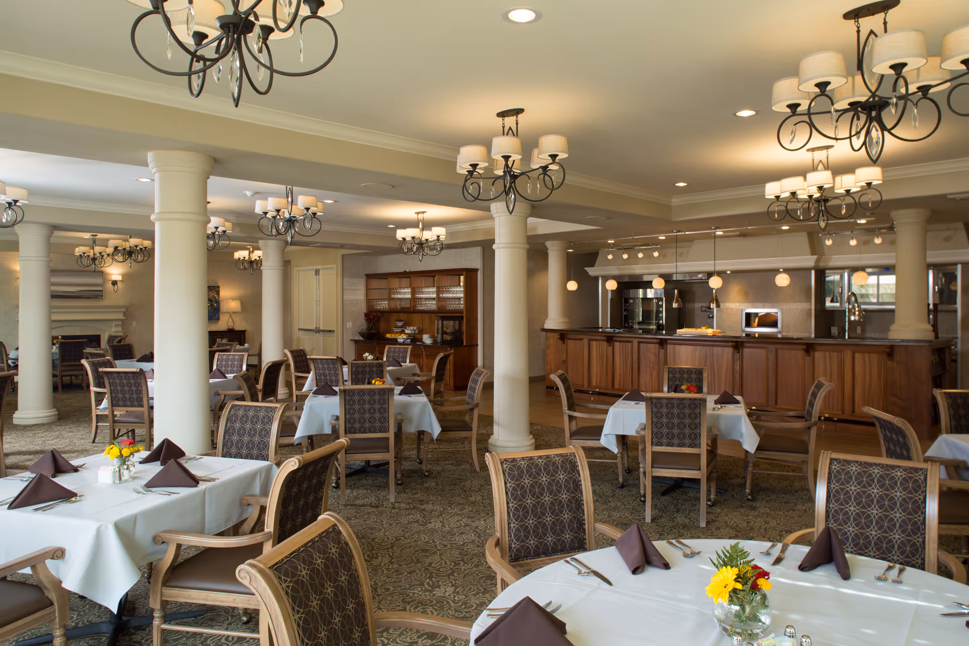 Elegant dining room with multiple set tables, upholstered chairs, chandeliers, columns, and a wooden service counter.