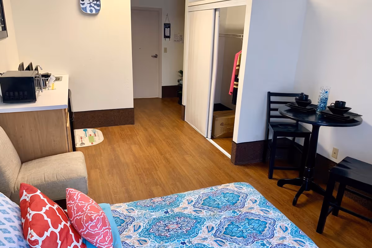 Interior of a senior living facility room with a bed covered in a blue and white patterned bedspread and red pillows, a small beige armchair, a kitchenette area with a microwave and sink, a small black dining table set with two chairs and black dishware, and an open closet with clothes and a cardboard box inside. The floor is wooden, and the walls are white with a brown baseboard.