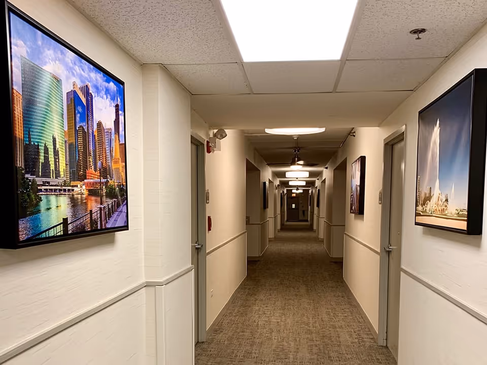 A long, well-lit hallway in a supportive living facility with beige walls and carpeted floor. Several closed doors line both sides of the corridor. Framed photographs of cityscapes and fountains hang on the walls. Ceiling lights and a ceiling fan provide illumination.