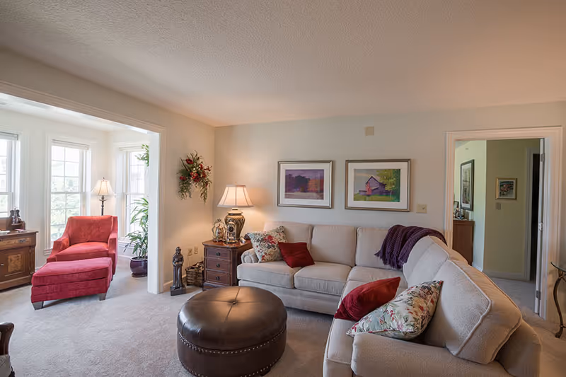 Well-lit living room with a beige sectional sofa, round leather ottoman, red armchair by bay windows, and lamps and framed artwork.