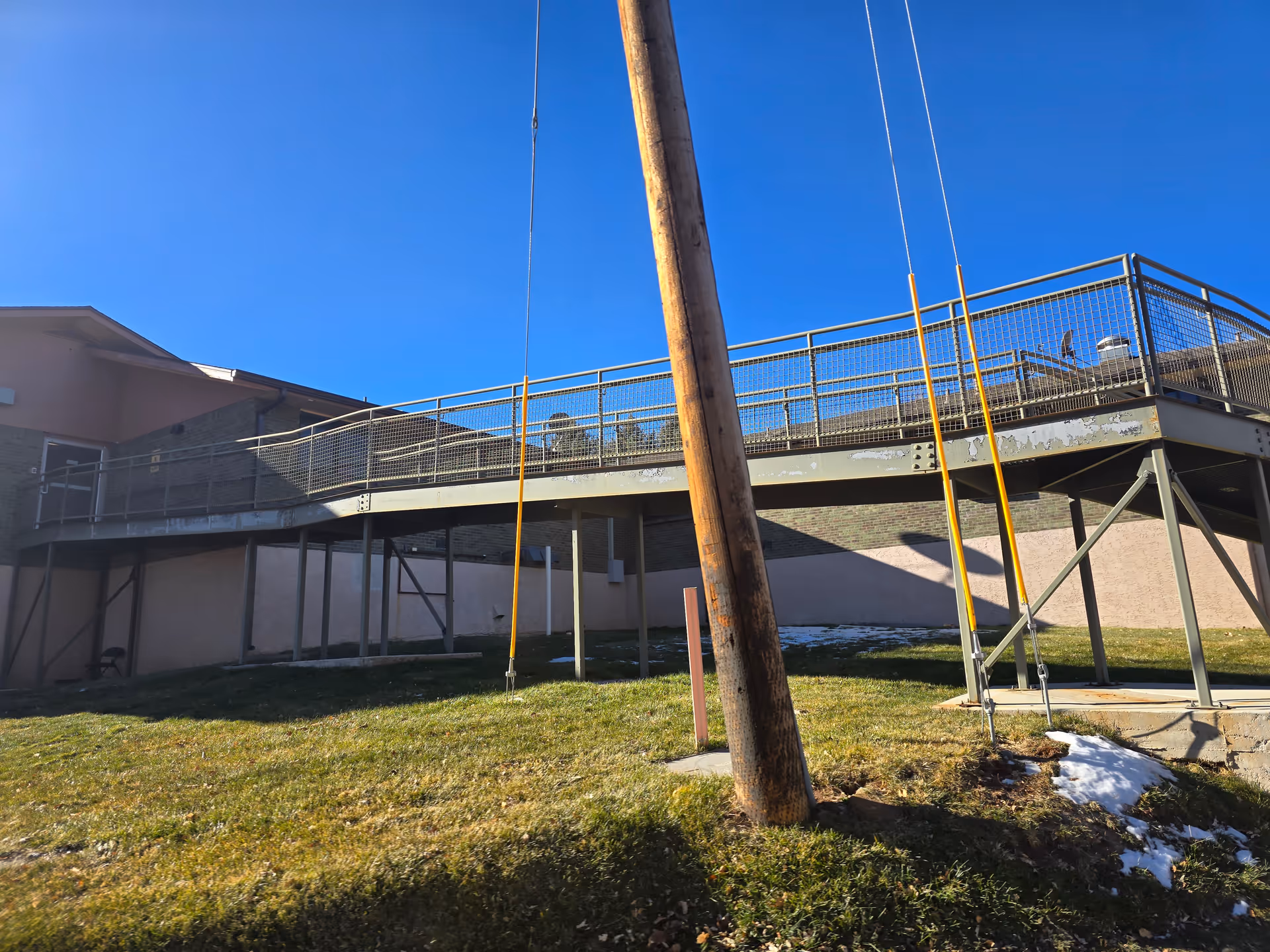 Outdoor view of a building with a raised metal walkway supported by metal beams. There is a wooden utility pole in the foreground and patches of snow on the grass below. The sky is clear and blue.