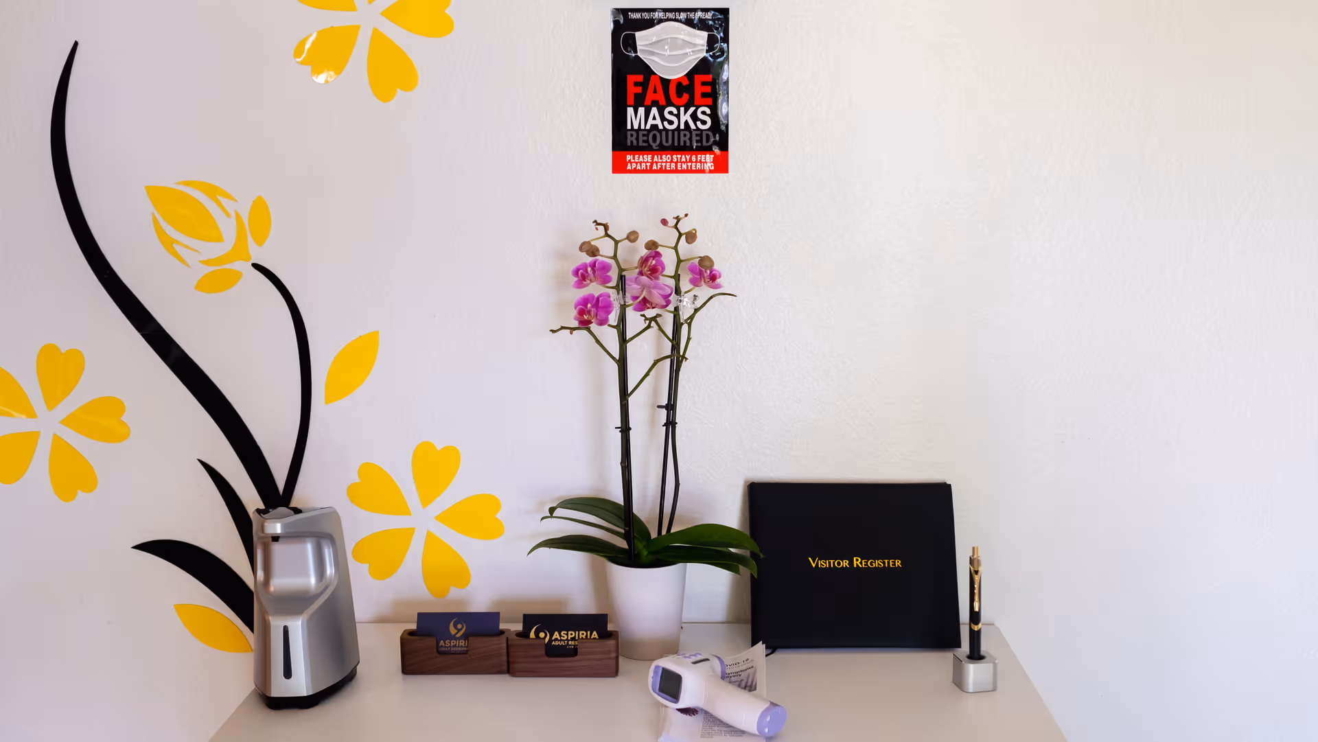 Reception desk area with a visitor register book, a pen in a holder, a digital thermometer, two business card holders with Aspira Adult Residences cards, a potted orchid plant with pink flowers, a hand sanitizer dispenser, and a wall decorated with yellow flower decals. A sign on the wall above reads 'FACE MASKS REQUIRED' with additional instructions.