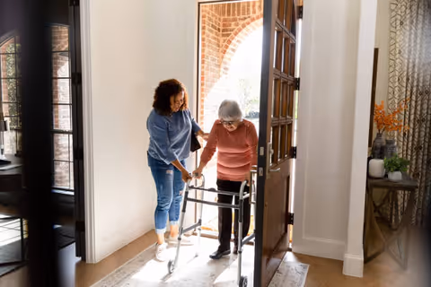 An elderly woman using a walker is being assisted by a younger woman as they enter through a wooden front door into a well-lit home interior with a rug and decorative items on a side table.