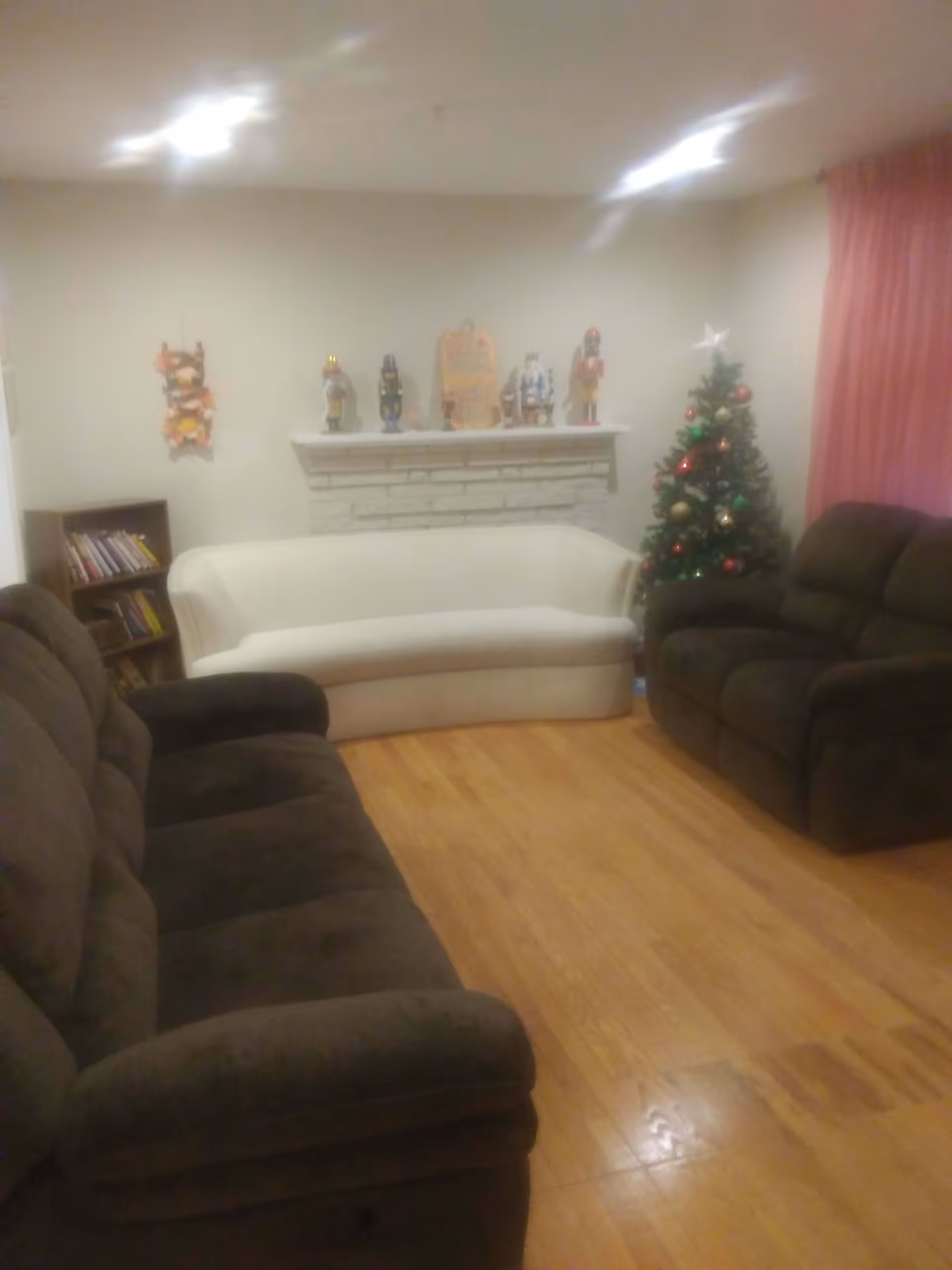 A cozy living room with two dark brown sofas and one white sofa arranged around a wooden floor. There is a decorated Christmas tree with ornaments and a star on top near a window with pink curtains. A white brick fireplace mantel is adorned with nutcracker figurines and a decorative plaque. A small bookshelf with books is visible in the corner.