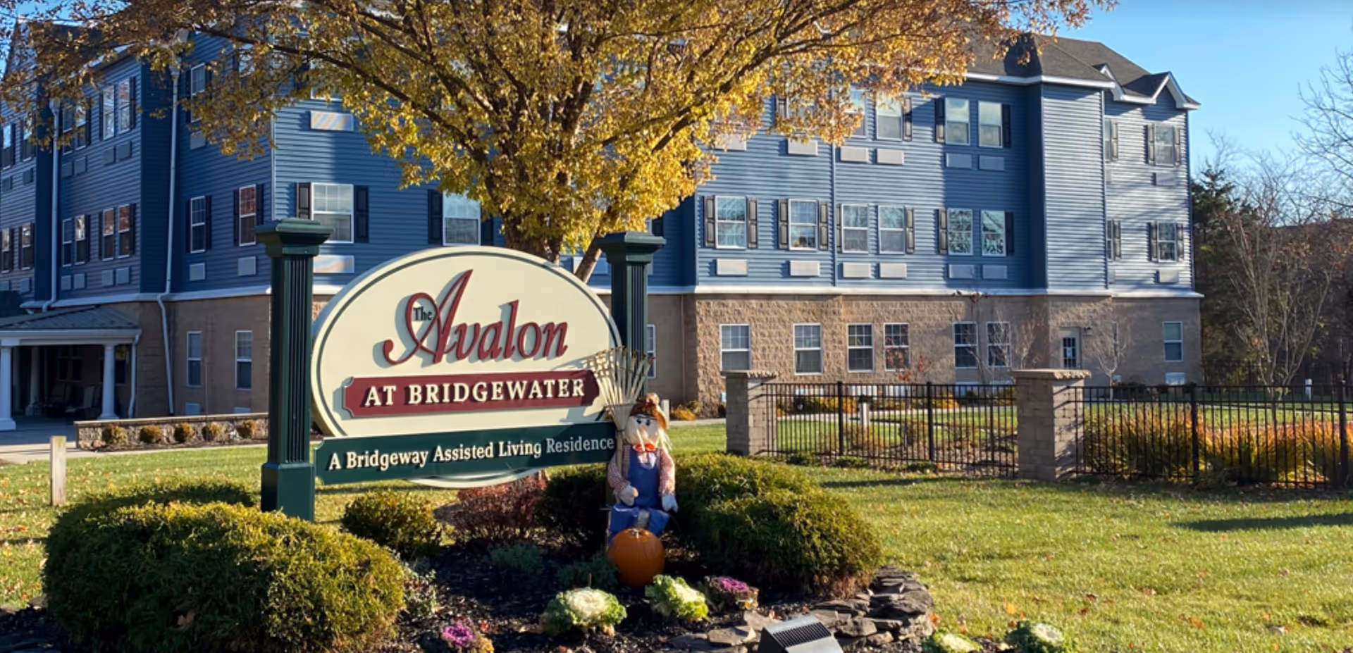 Exterior view of Avalon Assisted Living at Bridgewater, showing a multi-story building with blue siding and multiple windows. In front of the building is a landscaped area with bushes, flowers, and a sign that reads 'Avalon at Bridgewater, A Bridgeway Assisted Living Residence.' A scarecrow decoration and a pumpkin are placed near the sign, with a tree displaying autumn foliage in the background.