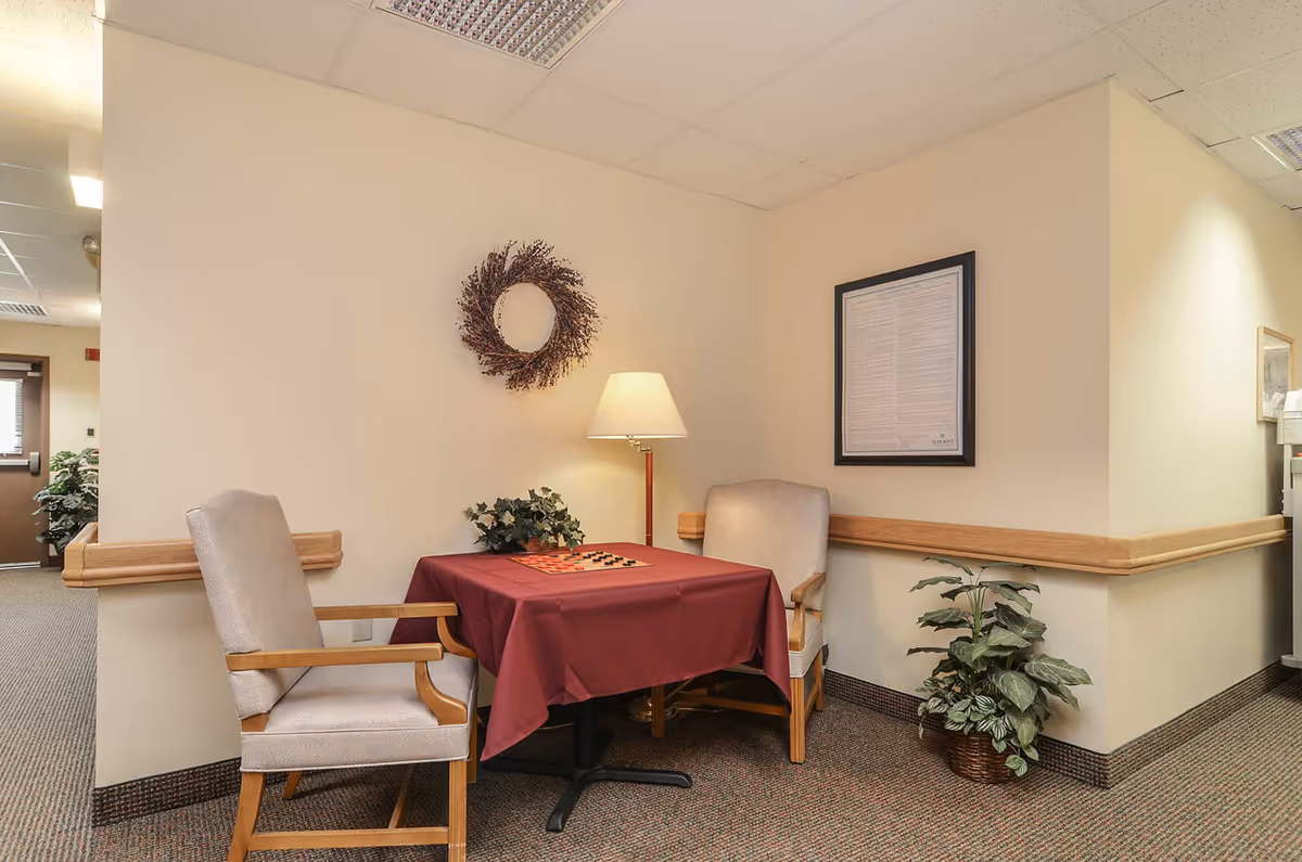 A small seating area in a senior living facility with two cushioned chairs around a table covered with a burgundy tablecloth. A checkerboard is on the table. The wall behind has a decorative wreath and a framed document. A floor lamp provides light, and there is a potted plant on the floor next to the table. The area has beige walls and carpeted flooring.