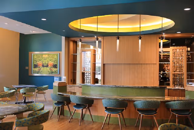Interior view of a modern dining area with a curved bar counter surrounded by green cushioned stools. The ceiling above the bar has a circular recessed lighting feature with hanging pendant lights. There are several tables and chairs with patterned upholstery, a large framed painting on the wall, and wooden flooring.