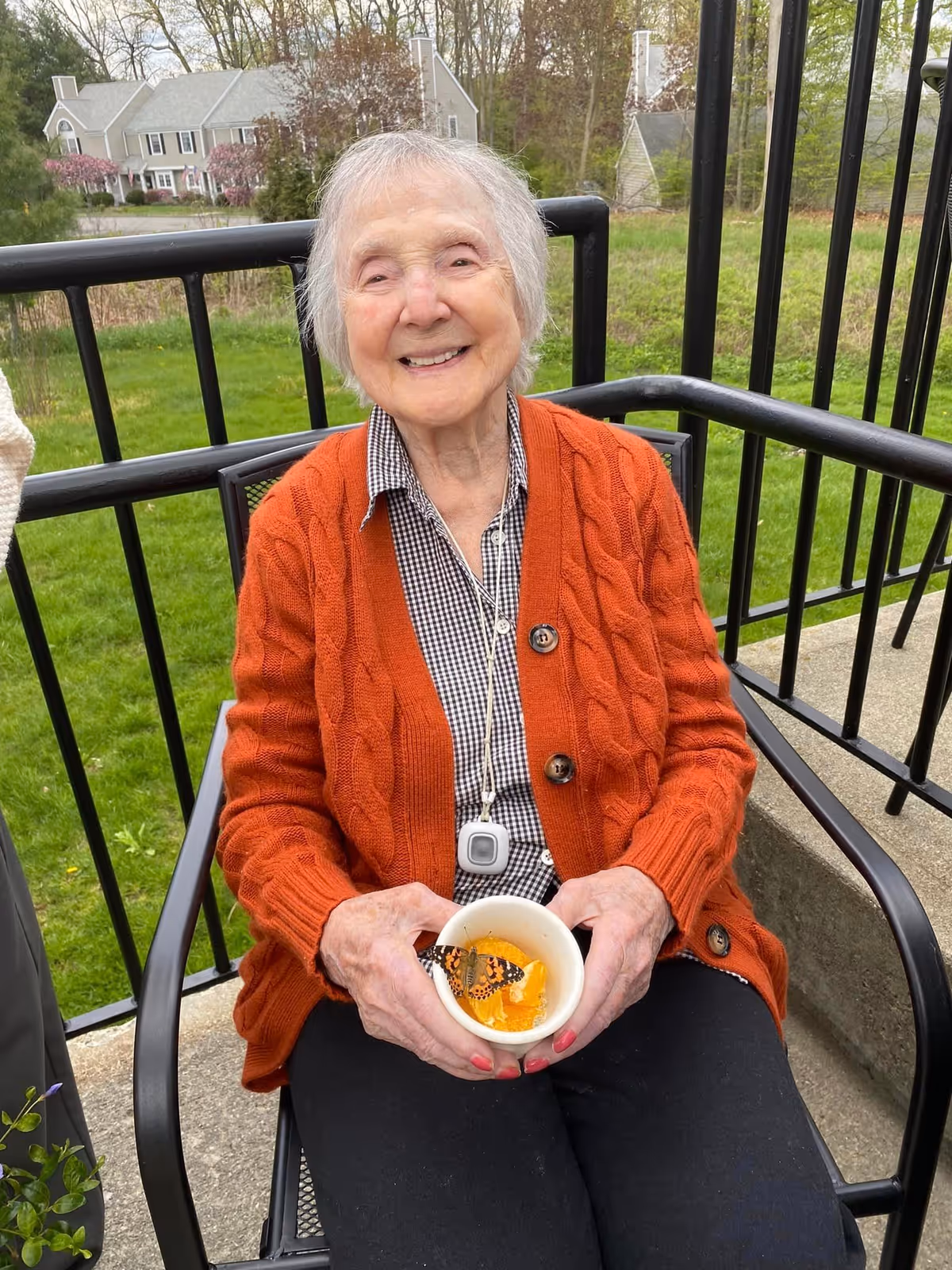 An elderly woman wearing an orange cardigan and a checkered shirt is sitting on a black metal chair outdoors on a patio. She is smiling and holding a white bowl with a butterfly perched on orange slices. Behind her is a black metal railing and a grassy area with trees and houses in the background.
