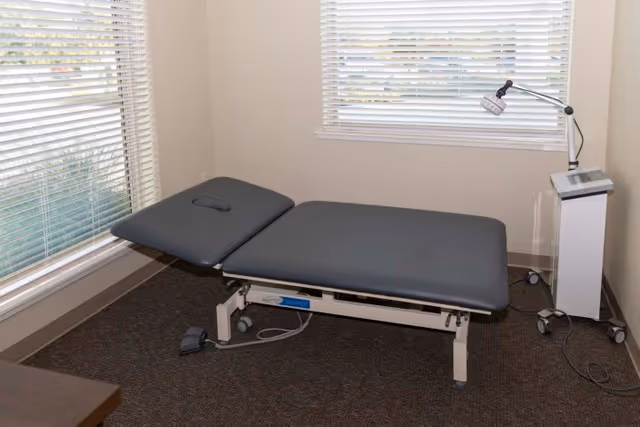 A medical examination or therapy room with a black adjustable treatment table positioned near two windows with white blinds. There is a white medical device with a lamp on an adjustable arm on the right side of the table. The room has beige walls and a dark carpeted floor.