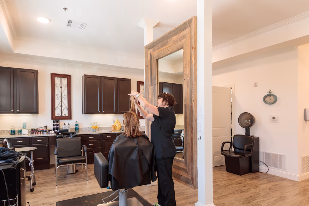 A hair salon area inside a senior living facility with a stylist cutting a seated woman's hair. The room features dark wood cabinets, a large rustic framed mirror, and salon chairs. The floor is wooden, and the walls are light-colored.