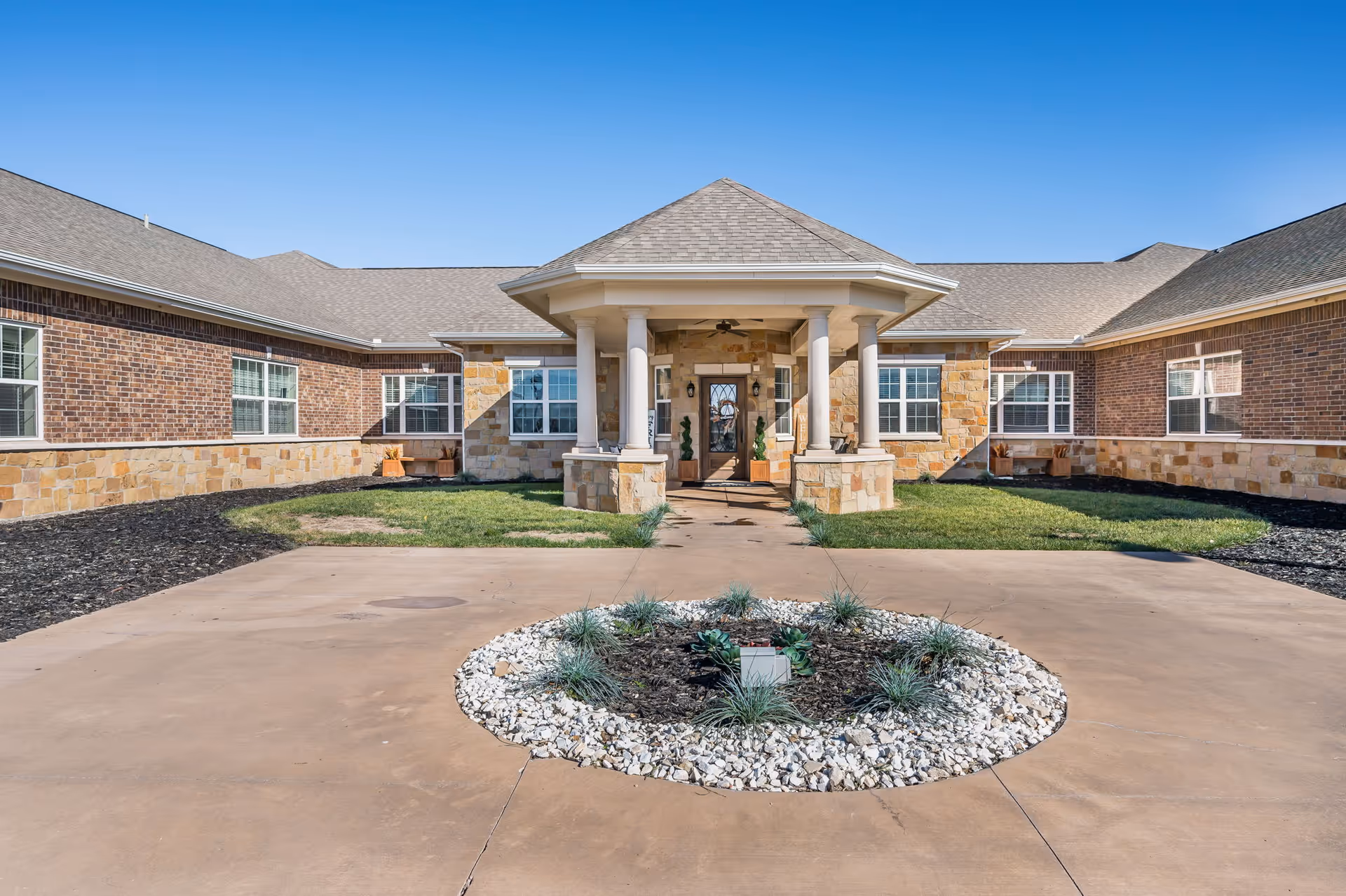 Front exterior view of The Gardens of Amarillo Memory Care facility with a circular driveway, landscaped center island with plants and white rocks, and a covered entrance supported by columns. The building features brick and stone facade under a clear blue sky.
