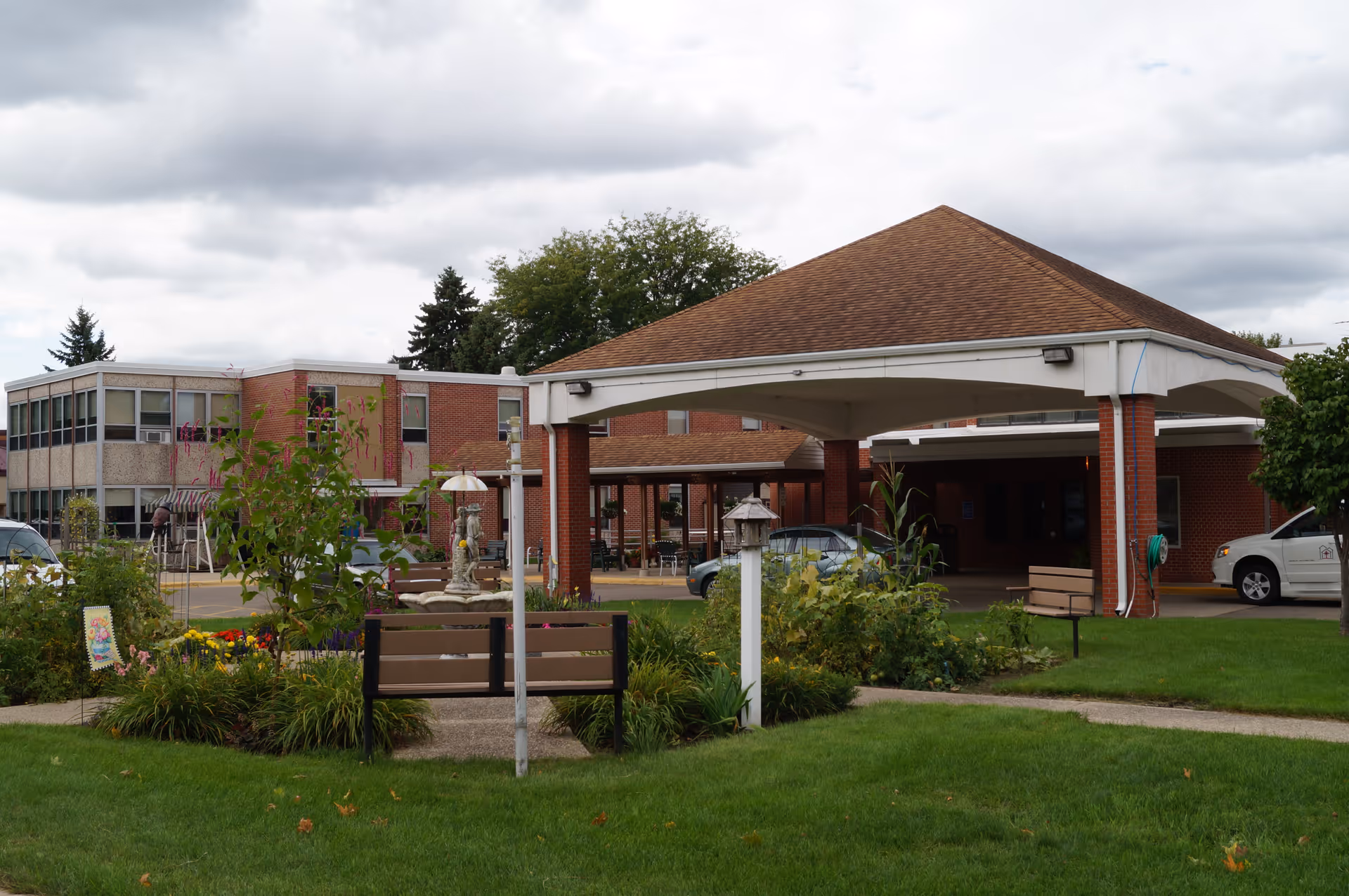Exterior view of St John's Lutheran Community showing a covered entrance with brick pillars, a garden area with benches and flowers in the foreground, and a two-story building in the background under a cloudy sky.