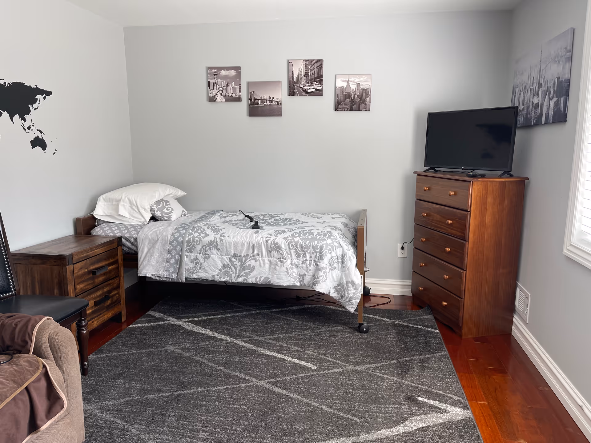 A small bedroom with a single bed covered in a gray and white patterned comforter and pillows. Next to the bed is a wooden nightstand and a black chair. On the right side of the room is a wooden dresser with a flat-screen TV on top. The walls are light gray with black and white cityscape pictures hanging above the bed and on the adjacent wall. A gray area rug with white lines covers part of the wooden floor.