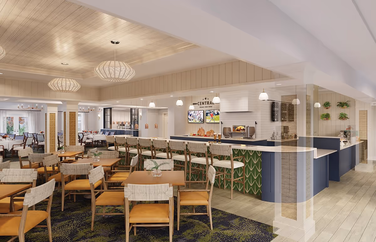 A spacious and modern dining area with multiple wooden tables and chairs featuring cushioned seats. There is a bar counter with high chairs and pendant lights hanging above. The ceiling has a wooden panel design with additional light fixtures. In the background, there are more dining tables with white tablecloths and a kitchen or serving area with two TV screens mounted on the wall. The decor includes plants on the walls and a mix of carpet and wooden flooring.