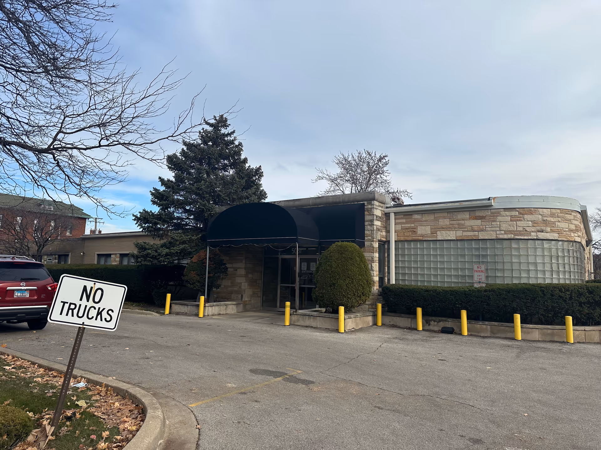 Entrance to a single-story rehab building with a black awning, curved glass-block wall, and a "NO TRUCKS" sign in the driveway.